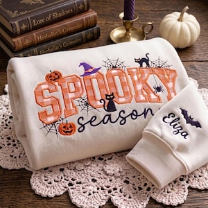 May include: A cream-coloured sweatshirt featuring the embroidered text SPOOKY season in orange plaid and purple script. The design includes pumpkins, a witch hat, black cats, and spiderwebs. The sleeve is embroidered with the name Eliza and a bat.