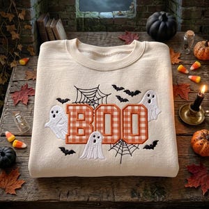 Könnte beinhalten: Ein cremefarbenes Sweatshirt mit Rundhalsausschnitt und einer Stickerei des Wortes BOO in orange-weiß karierten Blockbuchstaben, umgeben von weißen Geisterfiguren, schwarzen Spinnennetzen und kleinen schwarzen Fledermäusen auf der Vorderseite.