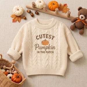 Puede incluir: Un suéter de punto trenzado color crema con el texto marrón "CUTEST Pumpkin IN THE PATCH" y un gráfico de calabaza naranja en el centro. El suéter tiene un cuello redondo acanalado, mangas largas con puños acanalados y una textura de punto grueso.