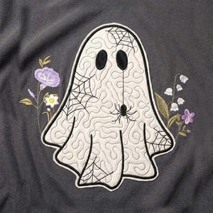 Puede incluir: Una sudadera gris carbón que presenta un gran fantasma blanco bordado con ojos ovalados negros, una araña negra y telarañas. El fantasma está rodeado de ramas florales bordadas con flores moradas y blancas a ambos lados.