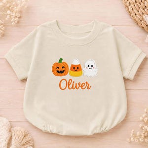 May include: A cream-colored short-sleeve romper featuring an embroidered design of a smiling jack-o'-lantern, a piece of candy corn, and a ghost. Below the icons, the name Oliver is embroidered in orange cursive script on the front.