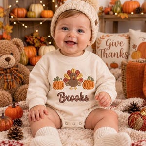 May include: A cream-colored long-sleeve bodysuit featuring an embroidered turkey flanked by two orange pumpkins and the name Brooks in brown script. The garment is worn with a matching cream-colored knit beanie and ribbed socks.