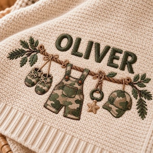 May include: A cream-coloured waffle-knit blanket featuring an embroidered design of a rope with green camouflage-patterned baby items including shoes, overalls, and a cap, along with a star and the name OLIVER stitched in dark green thread.
