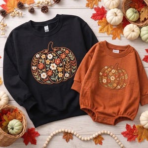 May include: A black long-sleeve sweatshirt and an orange long-sleeve romper, both featuring a central graphic of a pumpkin filled with various colorful flowers and leaves. The garments have ribbed crew necklines and cuffs, with a snap-button closure.