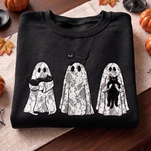 May include: A black crewneck sweatshirt featuring an embroidered design of three ghosts draped in white floral lace fabric. Each ghost is paired with a black cat, with one cat held in the arms, one perched on top, and one held vertically.
