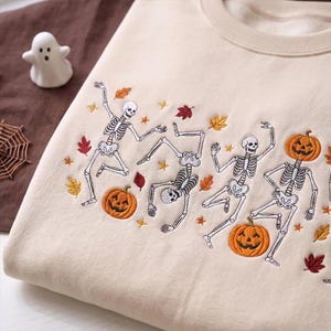 May include: A beige crewneck sweatshirt featuring an embroidered design of white skeletons dancing alongside orange jack-o'-lantern pumpkins and scattered autumn leaves in shades of red, yellow, and orange, accented with small gold star shapes.