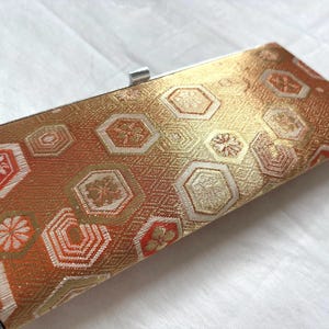 May include: A rectangular clutch bag featuring a metallic gold and burnt orange woven fabric with a repeating hexagonal pattern. The exterior surface displays intricate geometric shapes and floral motifs. A silver-toned metal frame and clasp are visible.