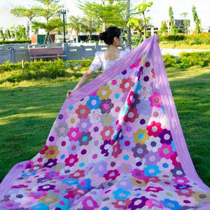 Può includere: Una grande trapunta fatta a mano con un motivo floreale nei toni del rosa, viola, blu e giallo. La trapunta è tenuta in alto, mostrando l'intricato design e l'artigianato. Il bordo è di colore lavanda.