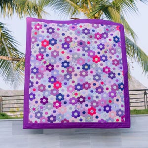 May include: A handmade quilt with a floral pattern in shades of purple, pink, and white. The quilt features a repeating design of hexagonal flower shapes, bordered by a solid purple edge. The quilt is hanging outdoors.