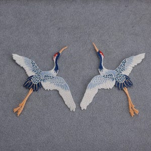 May include: Two embroidered crane patches. The cranes have white wings with blue accents, red crowns, and long, orange legs. The patches are on a gray background, and the cranes are facing each other.