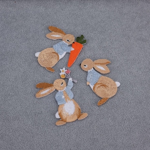 May include: Three embroidered bunny patches. Each bunny is brown with tan ears and a white belly. Two bunnies wear light blue jackets. One bunny holds a carrot, and another holds a bouquet of flowers. The patches are on a grey background.