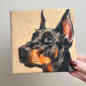 以下が含まれることがあります： ドーベルマン犬の正方形のキャンバス絵画。犬は横顔で描かれており、黒い毛並み、茶色のマーキング、警戒した表情をしています。背景は無地のベージュ色です。
