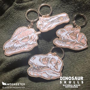 Op de afbeelding: Vier houten sleutelhangers met gedetailleerde illustraties van dinosaurus-schedels in wit- en lichtroze tinten. Elke sleutelhanger heeft een zilverkleurige metalen ring. De tekst DINOSAUR SKULLS NATURAL WOOD KEYCHAINS is zichtbaar in de hoek.