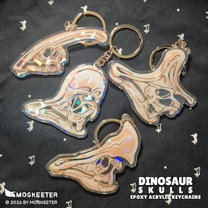 May include: Four acrylic keychains featuring detailed illustrations of dinosaur skulls with a holographic finish. Each keychain includes a gold-toned metal ring. The text DINOSAUR SKULLS EPOXY ACRYLIC KEYCHAINS, MOSKEETER, and 2026 BY MOSKEETER is visible.