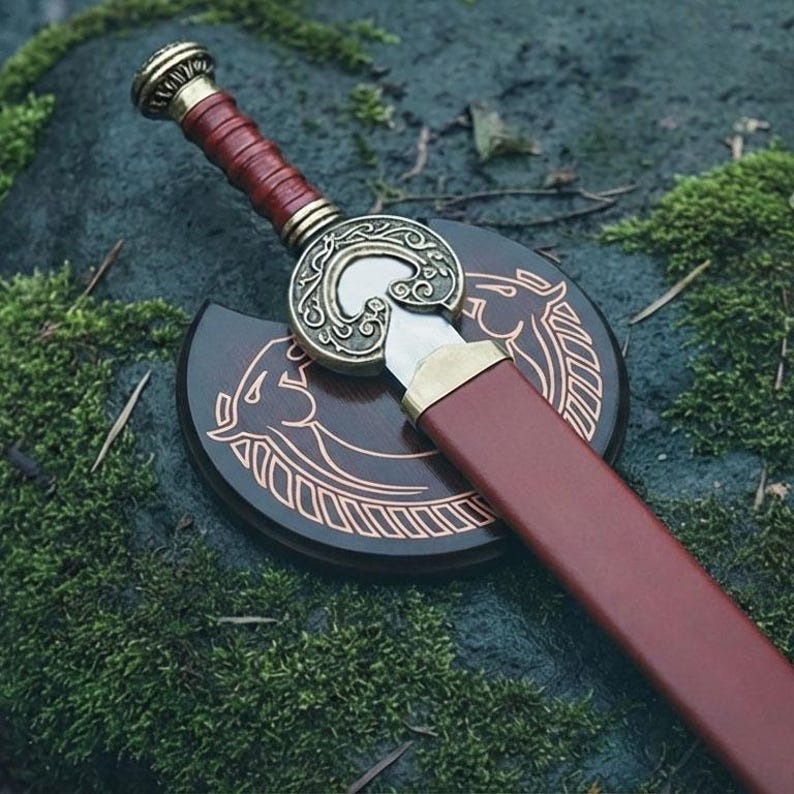 May include: A decorative sword with a brown leather-wrapped handle and a bronze-colored hilt. The sword rests on a wooden plaque with a horse design. The scabbard is a matching brown color. The sword is displayed on a mossy surface.