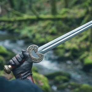 May include: A silver sword with an ornate, gold-colored hilt featuring a heart-shaped design. The sword's handle is wrapped in red leather, and the pommel is gold. The sword is held by a person wearing a black glove, with a forest background.