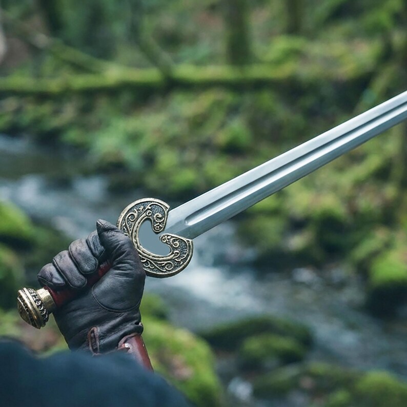 May include: A silver sword with an ornate, gold-colored hilt featuring a heart-shaped design. The sword's handle is wrapped in red leather, and the pommel is gold. The sword is held by a person wearing a black glove, with a forest background.