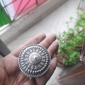 May include: A round, silver-colored trinket box with an intricate, embossed floral design. The box is held in an open hand, showcasing its detailed craftsmanship. The box has a hinged lid and a small clasp.