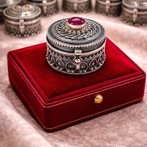 May include: A silver-toned, round jewelry box with ornate detailing and a red gemstone on the lid, resting on a red velvet box. Several similar boxes are visible in the background, suggesting a collection or set.