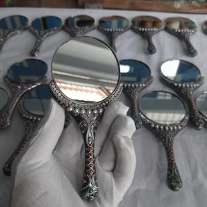 May include: Handheld silver mirrors with round reflective surfaces. The mirrors have ornate handles decorated with red and green stones and white pearls. The mirrors are arranged on a white surface.