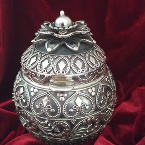 May include: Ornate silver-colored container with a domed lid and intricate floral and geometric patterns. The container has a small handle and sits on a red velvet surface. The lid is topped with a small, round finial.