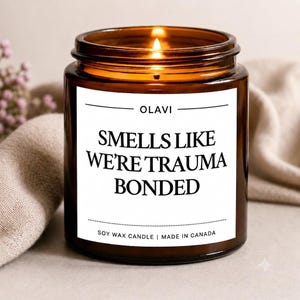 Peut inclure: Une bougie en cire de soja allumée dans un pot en verre brun avec une étiquette blanche. L'étiquette porte le texte OLAVI, SMELLS LIKE WE'RE TRAUMA BONDED, et SOY WAX CANDLE | MADE IN CANADA en polices noires avec des lignes horizontales.