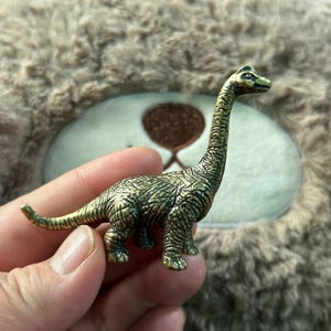 以下が含まれることがあります： 長い首と尾を持つ、小さく詳細な真鍮色の恐竜の置物。 恐竜は質感のある肌をしており、立っています。 置物の長さは約5cmで、背景はぼやけています。