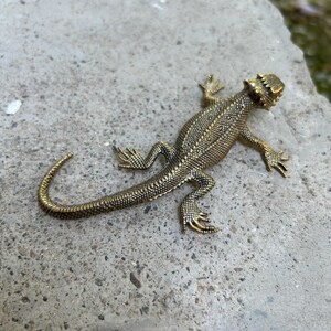 May include: A metallic gold-toned lizard figurine featuring a textured, scaly surface across its back, head, and long tail. The lizard is depicted in a crawling position with four legs extended outward, showcasing detailed skin patterns and a distinct head shape.