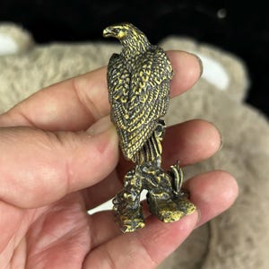 May include: A detailed brass eagle figurine with textured feathers and a detailed perch. The eagle is depicted in a standing position, with its head turned slightly to the side. The brass has a golden hue, and the figurine is likely a decorative item.