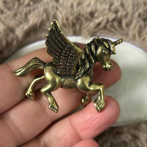 May include: A small, metallic brass-colored figurine of a winged unicorn in a galloping pose. The creature features detailed feathers on its wings, a flowing mane, and a single horn on its forehead, with a polished finish and dark accents.