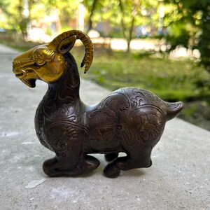 Könnte beinhalten: Eine bronzefarbene Ziegenfigur mit goldfarbenem Kopf und Hörnern. Die Ziege hat detaillierte Blumenmuster auf ihrem Körper. Die Figur steht.