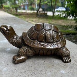 May include: A bronze-colored turtle figurine with detailed shell and textured skin. The sculpture is posed on a concrete surface, showcasing the intricate design of the turtle's shell and body. The figurine is a decorative item.