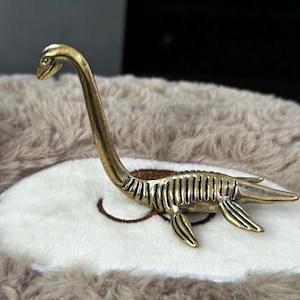 May include: A brass-toned sculpture of a plesiosaur, a marine reptile from the past, featuring a long neck and flippers. The sculpture has ribbed detailing along its body. The plesiosaur is on a white surface with a brown bear design.