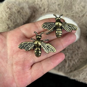 May include: Two antique brass bee-shaped decorative objects. The bees have detailed wings and body segments. The objects are likely used for jewelry, crafts, or home decor. The bees are held in a hand.