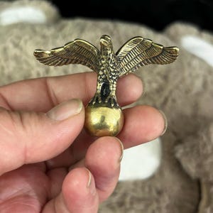 May include: A detailed brass eagle figurine with outstretched wings. The eagle is perched on a golden sphere. The wings and feathers are intricately detailed. The figurine is held in a person's hand.