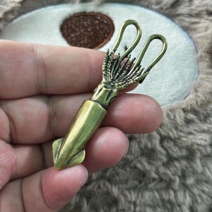 May include: A brass-toned metal squid figurine featuring a smooth, elongated mantle and detailed, textured tentacles at the top. The object has a metallic finish with subtle dark highlights in the crevices, showcasing a realistic marine creature form.