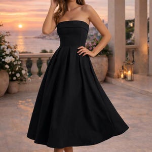 May include: A strapless black midi dress with a flared skirt. The dress is knee-length and paired with gold heels. The background features a sunset over the ocean and a stone balcony with white flowers.