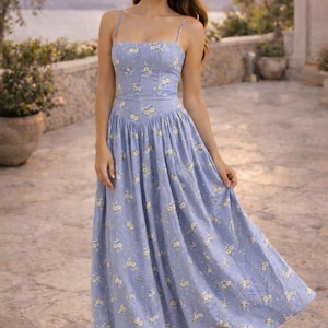 May include: A periwinkle blue midi dress featuring a square neckline, thin spaghetti straps, and a fitted bodice. The fabric is adorned with a small white and yellow floral pattern, and the skirt has a gathered waistline that flows into a full silhouette.