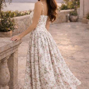 May include: A midi-length dress featuring a white base with a delicate pink and green floral print. The garment has thin spaghetti straps, a fitted bodice, and a full, pleated skirt that falls below the knees, crafted from a textured fabric.