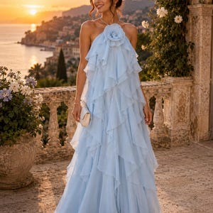 May include: A floor-length light blue halter dress featuring cascading vertical ruffles throughout the skirt and a large fabric flower accent at the neckline. The garment has a flowing silhouette and is paired with a small white clutch bag.