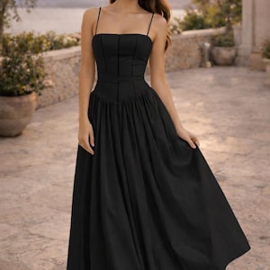 May include: A black midi dress featuring a fitted bodice with vertical seam detailing, a straight neckline, and thin spaghetti straps. The skirt is gathered at the waist, creating a full, flowing silhouette that falls below the knees.