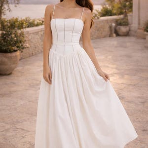 May include: A white midi dress featuring a fitted bodice with thin spaghetti straps and vertical seam detailing. The waistline is defined by a structured band, transitioning into a full, pleated skirt that falls to a mid-calf length.