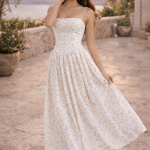 May include: A midi-length dress in a cream-colored fabric featuring a delicate, small-scale floral print in shades of yellow, blue, and pink. The garment has a fitted bodice with thin spaghetti straps, a straight neckline, and a full, flowing skirt.