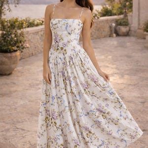 May include: A white midi dress featuring a delicate floral print in shades of purple, green, and blue. The garment has a fitted bodice with a straight neckline, thin spaghetti straps, and a full, pleated skirt that falls to a mid-calf length.