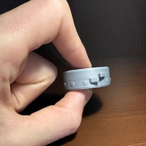 May include: A small, circular, grey 3D-printed object with a flat top and bottom. The side features a series of evenly spaced, raised circular studs and a small, protruding rectangular tab, giving it the appearance of a mechanical component.