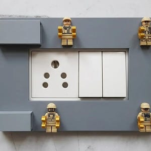 May include: A rectangular gray wall plate cover featuring a central white electrical outlet and two light switches. The frame is decorated with four small tan plastic toy figures positioned at the corners and two gray rectangular blocks.