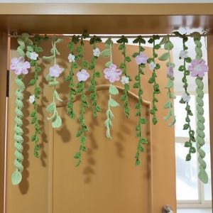 May include: A crocheted floral garland with green vines, leaves, and pink and white flowers hangs in front of a door. The garland is made of yarn and is designed to be decorative.