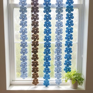 May include: Crocheted flower garlands in shades of blue and brown hang in front of a window. The garlands are made of individual flower shapes, creating a decorative and textured effect. A small potted plant sits on the windowsill.