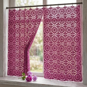 Könnte beinhalten: Ein magentafarbener Häkelvorhang mit einem sich wiederholenden quadratischen Blumenmuster. Der Vorhang hängt an einer dunklen Metallstange mit Ringen und ist teilweise zur Seite gerafft, wodurch ein Fensterrahmen und kleine lila Blumen sichtbar werden.