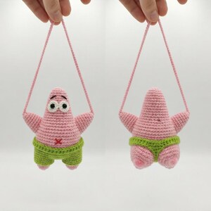 Puede incluir: Un peluche de ganchillo rosa con forma de estrella de mar, cuerpo estrellado, grandes ojos blancos con pupilas negras y pantalones cortos verdes. El juguete tiene una pequeña cruz roja bordada en la barriga y un cordón rosa para colgarlo.